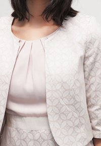 Blazer rose clair à motif avec une texture lisse et un col rond, associé à un haut plissé et une taille cintrée.