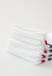 Chaussettes en coton blanc empilées soigneusement, avec une texture côtelée. Accentées de rayures rouges et navy en haut. Design doux et léger.