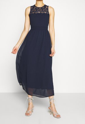 Femme portant une robe maxi bleu marine sans manches avec des détails en dentelle sur la poitrine et des sandales à talons hauts à brides gris clair.