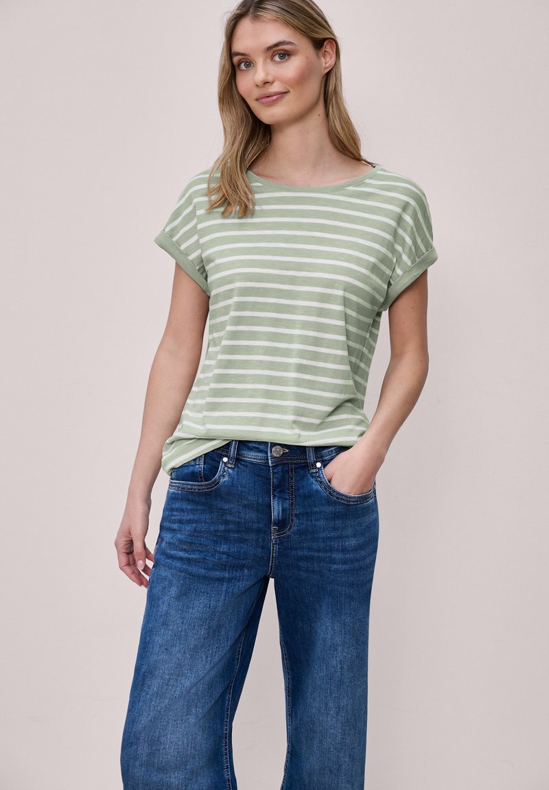 Femme debout, une main dans la poche, portant une chemise à manches courtes rayée vert clair et blanc, et un jean bleu, sur un fond neutre.