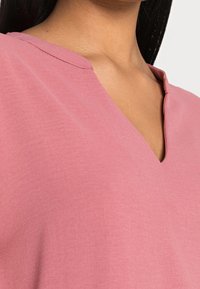 Rosa V-Ausschnitt-Oberteil mit einer weichen, glatten Textur; hat ein schlichtes Design und eine leicht lockere Passform. Detailreiche Nähte entlang des Ausschnitts.
