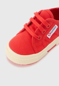 Scarpa da ginnastica rossa in tela con superficie texturizzata, suola in gomma bianca e lacci rossi. Presenta un logo blu e rosso sul lato. Design con punta arrotondata.