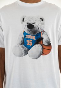 Camisa blanca con un gráfico de un oso de peluche gris vestido con una camiseta azul que lleva impreso "DUNK" y "35" en naranja, sosteniendo un baloncesto.