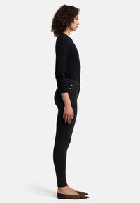 Jeans skinny noirs à taille haute, associés à des ballerines pointues au motif léopard. Cheveux bouclés, vue de côté, accessoires minimes.