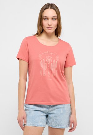 Woman with straight brown hair wears coral pink t-shirt with cactus print and light denim shorts, standing against plain background.