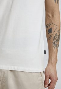 Bijela pamučna majica s zaobljenim rubom i božnim oznakom. Na modelovoj koži vidljiv je tetovaža karte koja se nalazi na ruci.