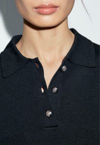 Polo shirt nero con texture, dotato di colletto, tre bottoni con dettagli metallici e una piccola tasca frontale.