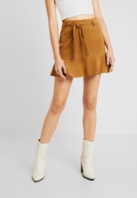 Jupe mini en peau suédée marron avec un ourlet évasé, une ceinture à nouer à la taille et une texture lisse. Complétée par des bottes blanches à talons.