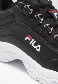 Zapatilla Fila negra con parte superior de cuero liso, suela de goma blanca y ojales de metal para los cordones. Presenta un logotipo bordado en blanco y rojo.