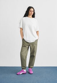 Λευκό υπερμεγέθες t-shirt, ελαιχ πράσινο παντελόνι με κορδόνι και ροζ αθλητικά παπούτσια με λευκές λεπτομέρειες. Το μοντέλο στέκεται μπροστά σε ουδέτερο φόντο.