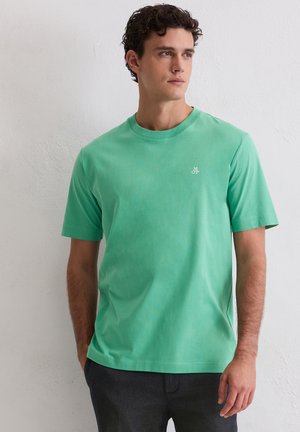 Młody mężczyzna z kręconymi włosami, ubrany w gładki turkusowy T-shirt z krótkim rękawem i małym białym logo "M OP" na piersi, stoi na tle białej ściany.