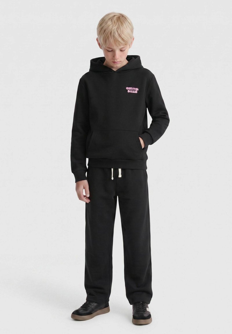 Enfant blond portant un sweat à capuche noir avec un texte rose et un pantalon de survêtement noir, debout, regardant vers le bas, une main dans la poche et une expression neutre.