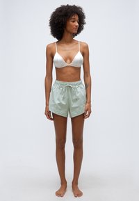 Γυναίκα με σγουρά μαλλιά, ντυμένη με λευκό σουτιέν και ανοιχτοπράσινο σορτς με κορδόνι, όρθια ξυπόλυτη μπροστά σε απλό ανοιχτό φόντο.