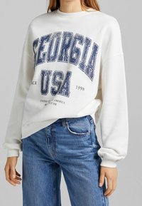 Sweat-shirt blanc arborant l'inscription "GEORGIA USA" en imprimé graphique bleu marine, avec des poignets côtelés et un col rond, associé à un jean bleu clair.
