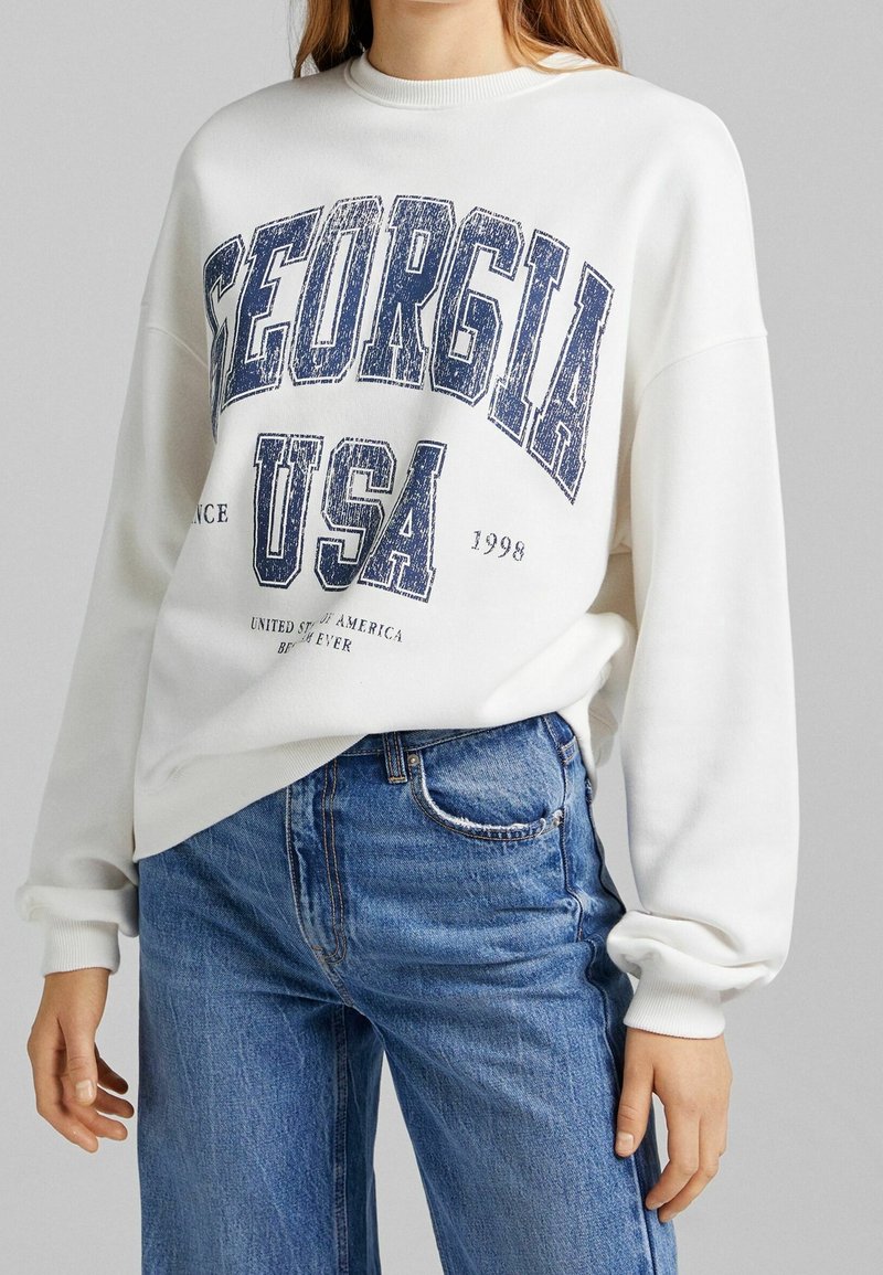 Sweat-shirt blanc arborant l'inscription "GEORGIA USA" en imprimé graphique bleu marine, avec des poignets côtelés et un col rond, associé à un jean bleu clair.