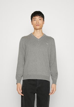Uomo che indossa un maglione grigio a scollo a V con maniche lunghe e pantaloni neri, in piedi davanti a uno sfondo chiaro e uniforme.