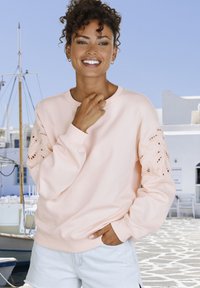 Lichtroze sweatshirt met decoratieve gaatjespatronen op de mouwen, een ronde halslijn en geribbelde manchetten; gecombineerd met lichte denim shorts.