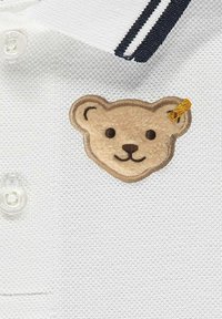 Weißes Poloshirt aus strukturierter Stoff mit einem Bärenkopf-Patch in Beige und Braun, akzentuiert mit einem kleinen goldenen Detail am Ohr.