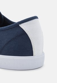 Bakhäl av en marinblå och vit kanvas sneakers med texturerad vit gummisula på en vit bakgrund.