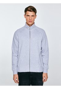 Młody mężczyzna w jasnoszarej kurtce zapinanej na zamek z długimi rękawami i czarnych spodniach, stojący na jednolitym jasnym tle.