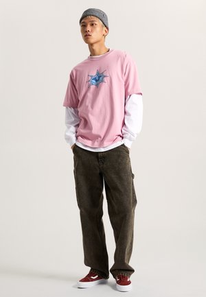Roze katoenen t-shirt met blauwe grafiek, gelaagd over een wit long-sleeve shirt; kaki cargobroek en rode suede sneakers maken de outfit af.
