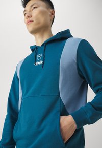 Felpa con cappuccio turchese con pannelli laterali azzurri, polsini a costine e tasca a marsupio; presenta il logo sul petto. Tessuto morbido.