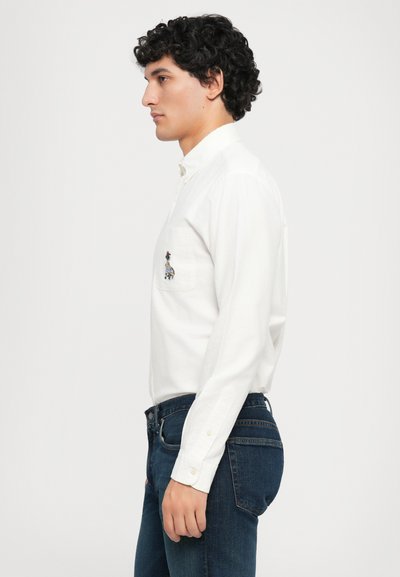 Cămașă albă cu mâneci lungi și nasturi, având un buzunar cu un design colorat brodat, asortată cu blugi denim de culoare albastru închis.