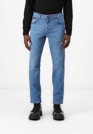 Jeans din denim de culoare albastră deschisă, cu tăietură dreaptă. Caracteristicile includ un design cu cinci buzunare, bucle pentru curea și un tiv cusut. Purtați cu cizme negre.