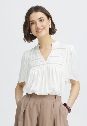 Mladá žena s krátkymi hnedými vlasmi sa usmieva, má na sebe bielu blúzku s krátkym rukávom a béžové nohavice s vysokým pásom, ruky v vreckách.