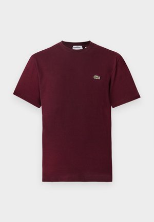 Mand og kvinde står side om side iført matchende mørkerøde t-shirts, lyse beige bukser og hvide sneakers foran en ensartet baggrund.