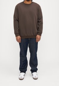 Brązowa bluza z okrągłym dekoltem i długimi rękawami, zestawiona z ciemnoniebieskimi jeansami i białymi trampkami. Miękki materiał, luźny krój, ściągacze na mankietach i u dołu.