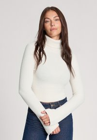 Maglione a collo alto bianco realizzato in un morbido tessuto texturizzato, con polsini e orlo a costine, abbinato a jeans aderenti blu scuro.