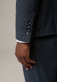 Giacca da suit blu scuro con una design aderente. Quattro bottoni neri decorano il polsino, in contrasto con un polsino di camicia bianco che sbuca.