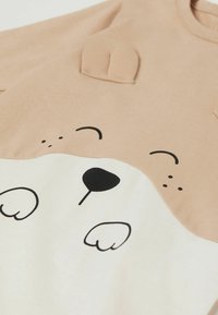 Beige Baumwoll-Sweatshirt mit einem weißen Bärenkopf-Design, das schwarze Druckakzente und weiche, gestickte Ohren-Details auf den Schultern aufweist.