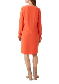 Oranges langärmliges Kleid mit gerader Silhouette und quadratischem Ausschnitt. Weicher, strukturierter Stoff, Rückenschlitz mit Knopfdetail, kombiniert mit beigen Blockabsatz-Stiefeln.
