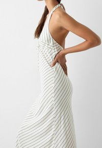 Randig halterneckklänning i krämfärg med tunna svarta vertikala linjer. Har en låg rygg och en samlad frontdetalj med knytning.
