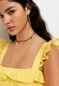 Mujer con el cabello largo y oscuro lleva grandes pendientes de aro dorados y collares dorados en capas, vestida con una blusa sin mangas amarilla con volantes.