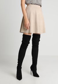 Falda de ante sintético beige con diseño acampanado, combinada con botas negras de textura hasta la rodilla con punta afilada y tacones de aguja.