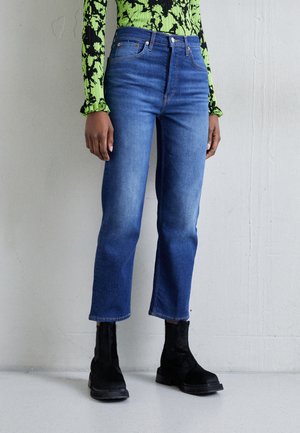 Osoba nosząca niebieskie krótkie spodnie jeansowe o prostych nogawkach, czarne botki za kostkę oraz zieloną, wzorzystą bluzkę z długim rękawem na tle jednolitej ściany.