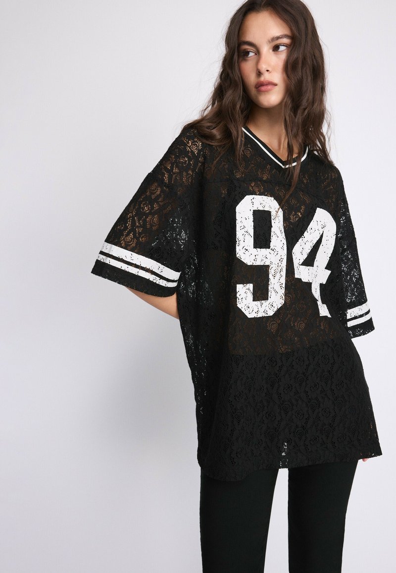 Jeune femme aux longs cheveux ondulés portant un top noir en dentelle oversize avec le numéro blanc 94 et des manches rayées, debout devant un fond clair uni.
