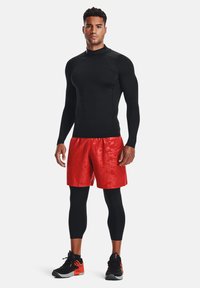 Haut de sport noir à manches longues, shorts rouges avec une texture brillante, leggings noirs et baskets noires avec des accents orange.