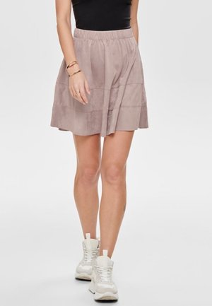 Femme portant une jupe en daim marron clair, un top noir sans manches, des baskets beiges et des bracelets dorés, debout devant un fond uni.