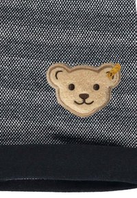 Pletený svetr s tmavě šedým a bílým vzorem, který má hnědý nášivku ve tvaru medvědí hlavy a malou žlutou stužku jako doplněk. Černý žebrovaný lem.