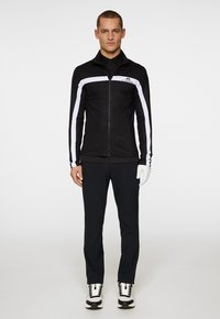 Black zip-up jacket with white side stripes, textured fabric; paired with black pants and white sneakers. Model wearing a glove.