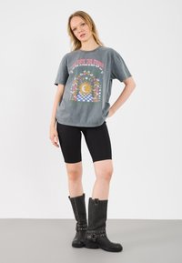 Harmaa graafinen t-paita, jossa on auringonkuva ja kukkakoristelut, yhdistettynä mustiin ribbatut pyöräilyshortseihin ja mustiin polveen yltäviin saappaisiin, joissa on metallikoristuksia.