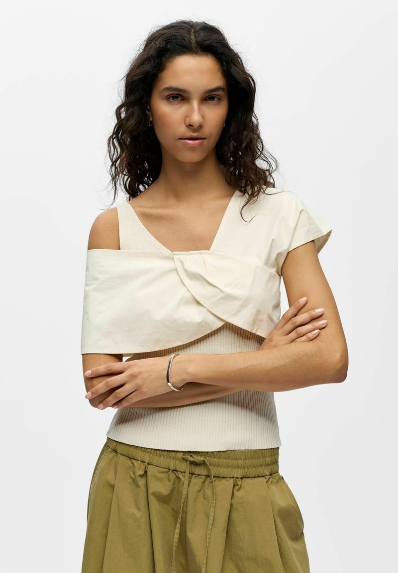 Femme aux cheveux bouclés foncés portant un haut écru avec un détail plié et une jupe vert olive, debout les bras croisés sur fond blanc.