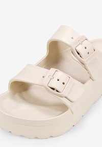 Sandales beige clair en matériau synthétique texturé avec deux sangles réglables dotées de boucles rectangulaires et d'une semelle rembourrée.