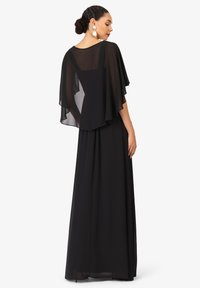 Schwarzes Maxikleid mit einem durchsichtigen, capeartigen Overlay. Glatter Stoff, lange Ärmel und ein taillierter Oberteil, der in einen locker fallenden Rock übergeht.