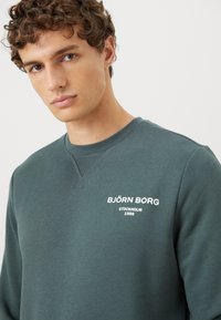 Nuori mies, jolla on kihara ruskea hius ja vihreä pyöreäkauluksinen collegepaita, jossa on valkoiset "BJÖRN BORG STOCKHOLM 1956" -tekstit rinnassa.