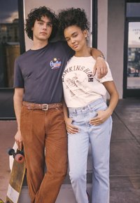 Young man holding skateboard, arm around young woman, both leaning against a pillar outside a building, wearing casual t-shirts and jeans.